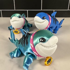 Puede incluir: Tres figuritas de tiburón impresas en 3D con un esquema de color azul, verde azulado y turquesa. Cada tiburón tiene un collar rosa con un patrón de huesos blancos y una etiqueta dorada que dice "Jeff".