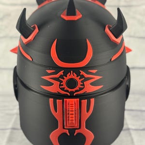Op de afbeelding: Een zwart en rode decoratieve helm met een gedetailleerd ontwerp. De helm heeft meerdere hoorns en spikes, met rode accenten en een zon-achtig embleem aan de voorkant. Het ontwerp is symmetrisch en gedetailleerd.