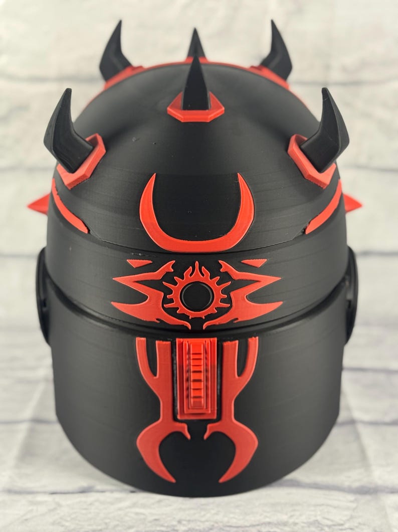 Op de afbeelding: Een zwart en rode decoratieve helm met een gedetailleerd ontwerp. De helm heeft meerdere hoorns en spikes, met rode accenten en een zon-achtig embleem aan de voorkant. Het ontwerp is symmetrisch en gedetailleerd.