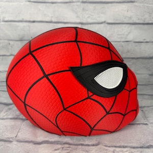 May include: A red Spider-Man mask featuring a textured surface with black web-like lines and large, angular black-rimmed eye lenses with white mesh inserts. The mask is designed with a rounded shape and displays a detailed, repeating pattern.