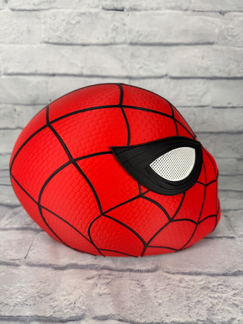 May include: A red Spider-Man mask featuring a textured surface with black web-like lines and large, angular black-rimmed eye lenses with white mesh inserts. The mask is designed with a rounded shape and displays a detailed, repeating pattern.