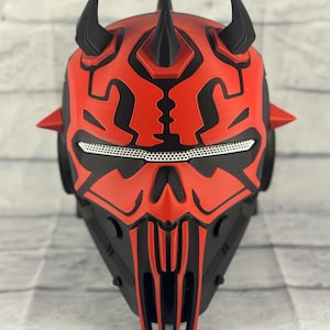 Op de afbeelding: Een rood en zwart masker met een gehoornd ontwerp, dat doet denken aan een personage uit een sciencefictionfilm. Het masker heeft ingewikkelde patronen, een mesh oogbedekking en een gedetailleerde, hoekige vorm. Het masker is gemaakt van een hard materiaal.