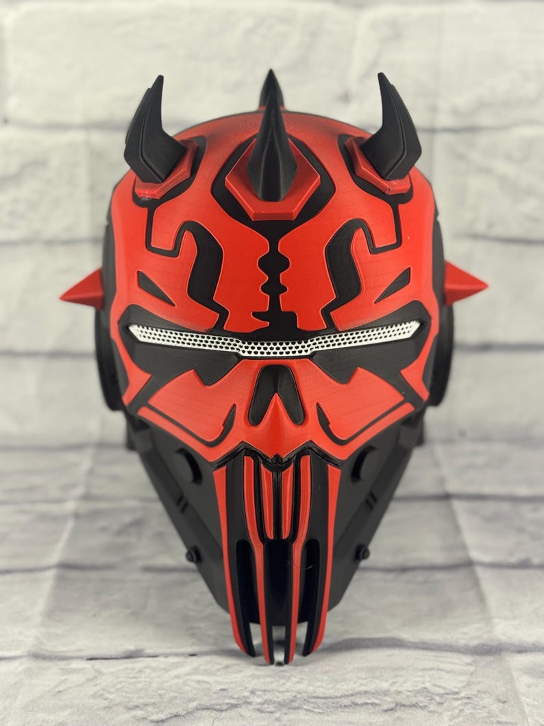 Op de afbeelding: Een rood en zwart masker met een gehoornd ontwerp, dat doet denken aan een personage uit een sciencefictionfilm. Het masker heeft ingewikkelde patronen, een mesh oogbedekking en een gedetailleerde, hoekige vorm. Het masker is gemaakt van een hard materiaal.