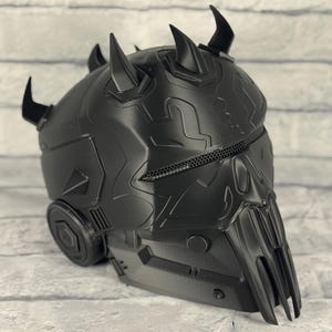 Op de afbeelding: Zwarte helm met een gedetailleerd, getextureerd oppervlak. De helm heeft meerdere hoorns en een gezichtsbescherming met een gaasventilatie. Het ontwerp heeft een futuristische, mechanische uitstraling.