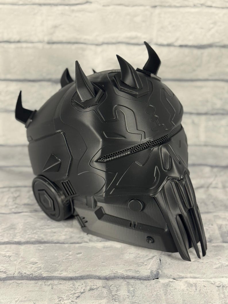 Op de afbeelding: Zwarte helm met een gedetailleerd, getextureerd oppervlak. De helm heeft meerdere hoorns en een gezichtsbescherming met een gaasventilatie. Het ontwerp heeft een futuristische, mechanische uitstraling.