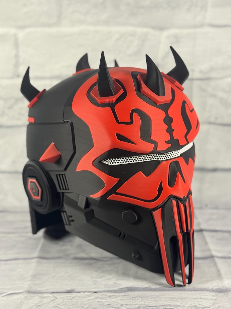 Op de afbeelding: Een zwart en rode helm met meerdere hoorns en een gedetailleerd ontwerp. De helm heeft een gaasgebied voor de mond en een reeks hoekige vormen. Het kleurenschema is voornamelijk zwart met rode accenten.