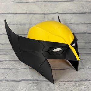 May include: A yellow and black superhero mask featuring a textured dome, pointed side fins, and white mesh eye coverings. The design includes a central yellow stripe running down the nose bridge, with a matte black finish on the lower face section.
