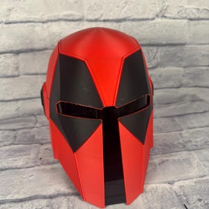 以下が含まれることがあります： 未来的なデザインの赤と黒のヘルメット。ヘルメットは、顔と側面に角張った黒のアクセントが付いた、滑らかで光沢のある赤い仕上がりになっています。デザインは、SFやコスプレ衣装を彷彿とさせます。