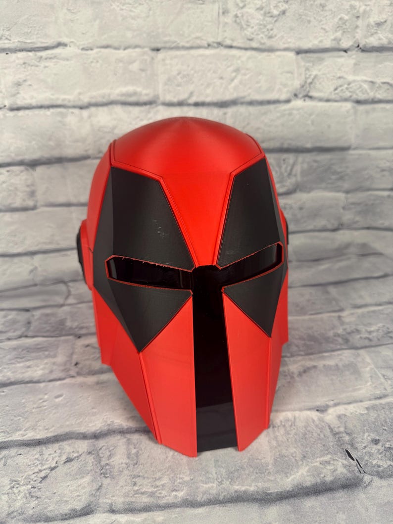 以下が含まれることがあります： 未来的なデザインの赤と黒のヘルメット。ヘルメットは、顔と側面に角張った黒のアクセントが付いた、滑らかで光沢のある赤い仕上がりになっています。デザインは、SFやコスプレ衣装を彷彿とさせます。