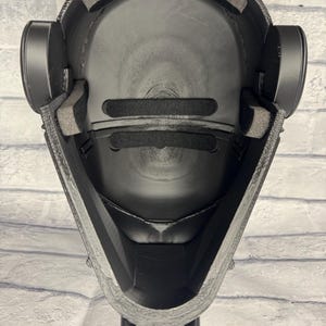 Op de afbeelding: Zwart helminterieur met gewatteerde voering en oordopjes. De helm heeft een getextureerde buitenkant en een gladde, afgeronde binnenkant. Het ontwerp omvat een kinbeschermer en een voorhoofdkussen. De helm is ontworpen voor bescherming en comfort.