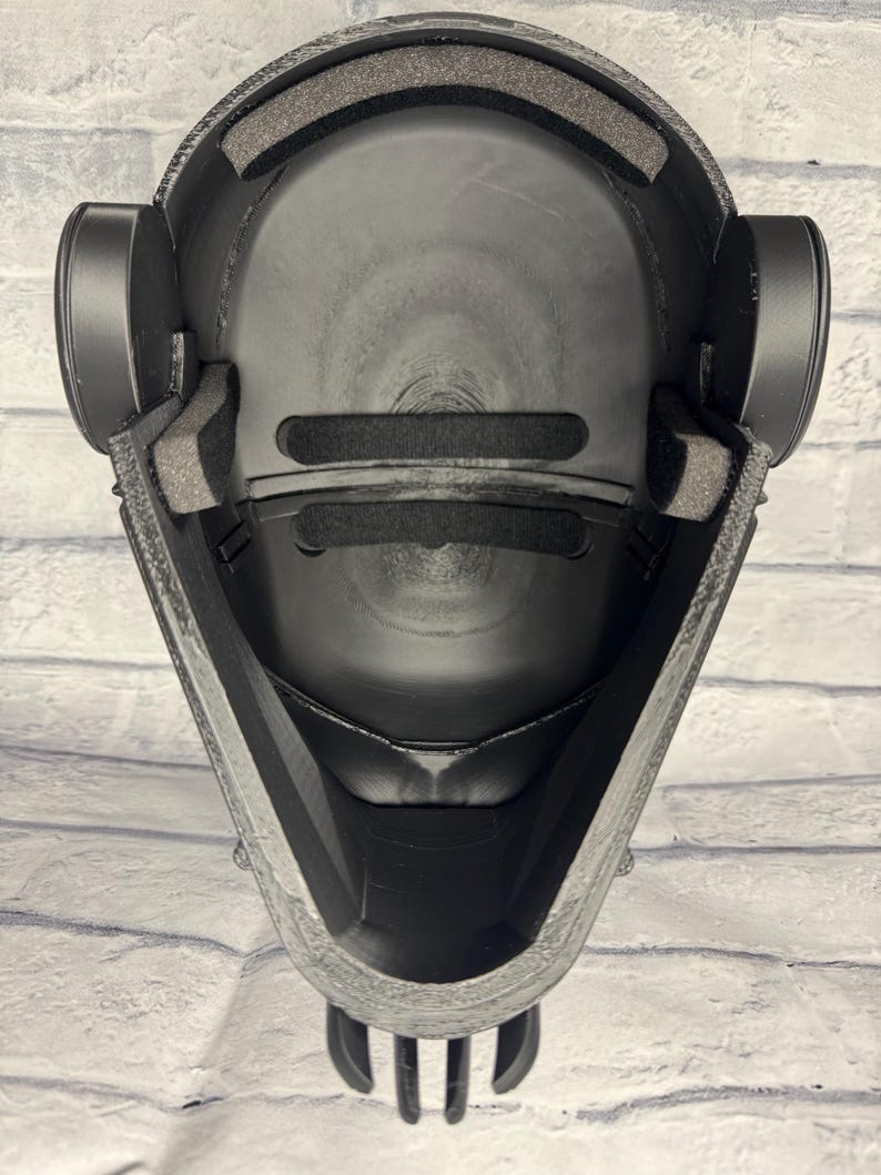 Op de afbeelding: Zwart helminterieur met gewatteerde voering en oordopjes. De helm heeft een getextureerde buitenkant en een gladde, afgeronde binnenkant. Het ontwerp omvat een kinbeschermer en een voorhoofdkussen. De helm is ontworpen voor bescherming en comfort.
