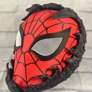 Puede incluir: Una m&aacute;scara roja y negra impresa en 3D con un dise&ntilde;o de telara&ntilde;a y zarcillos negros alrededor de los bordes. La m&aacute;scara tiene una cubierta de ojo de malla blanca.