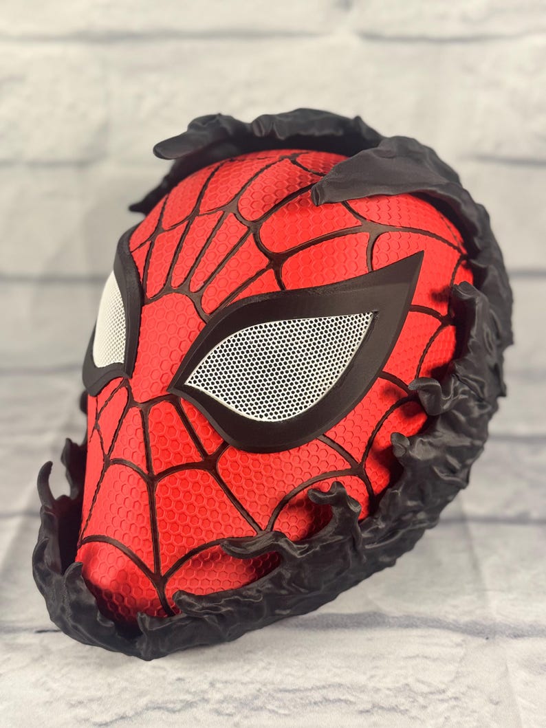 Puede incluir: Una m&aacute;scara roja y negra impresa en 3D con un dise&ntilde;o de telara&ntilde;a y zarcillos negros alrededor de los bordes. La m&aacute;scara tiene una cubierta de ojo de malla blanca.