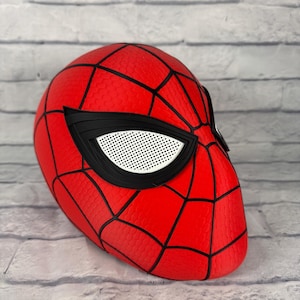 May include: A red Spider-Man mask featuring a black web pattern across the surface. The mask includes large, angular eye lenses with a white mesh texture framed by thick black borders, creating a distinct look for this superhero headgear.
