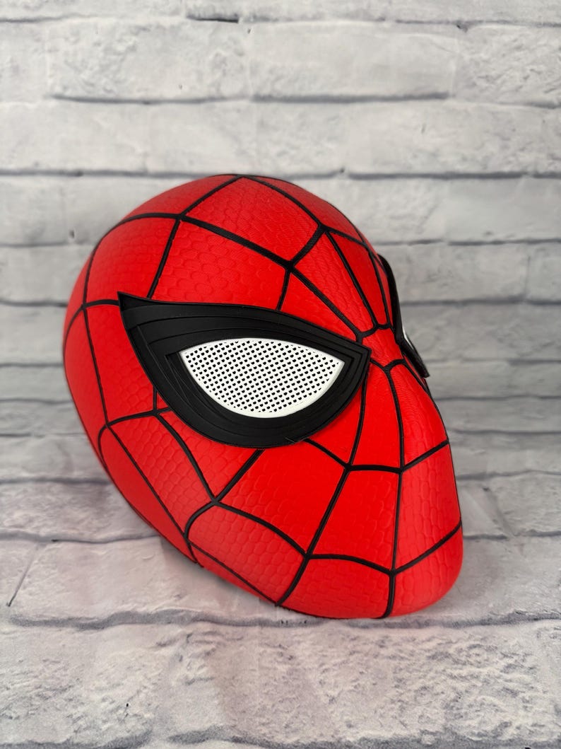 May include: A red Spider-Man mask featuring a black web pattern across the surface. The mask includes large, angular eye lenses with a white mesh texture framed by thick black borders, creating a distinct look for this superhero headgear.