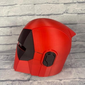 以下が含まれることがあります： 赤と黒のヘルメット。洗練された、角張ったデザインです。ヘルメットにはバイザーとイヤーピースが付いています。滑らかな素材で作られており、コスプレやコスチュームの一部である可能性があります。