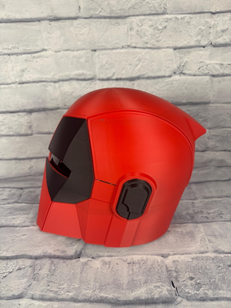 以下が含まれることがあります： 赤と黒のヘルメット。洗練された、角張ったデザインです。ヘルメットにはバイザーとイヤーピースが付いています。滑らかな素材で作られており、コスプレやコスチュームの一部である可能性があります。