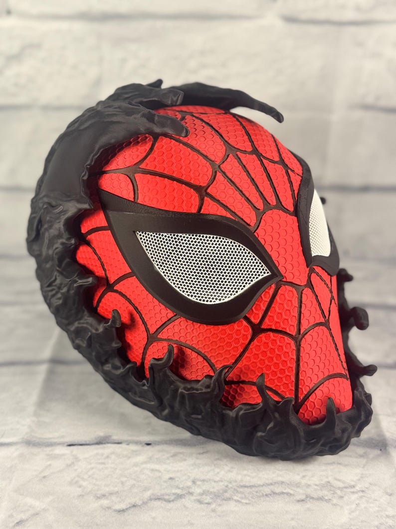 Puede incluir: Una m&aacute;scara impresa en 3D roja y negra con un dise&ntilde;o de telara&ntilde;a negra y un dise&ntilde;o de ara&ntilde;a negro texturizado alrededor de los bordes. La m&aacute;scara tiene una cubierta de ojo de malla blanca.