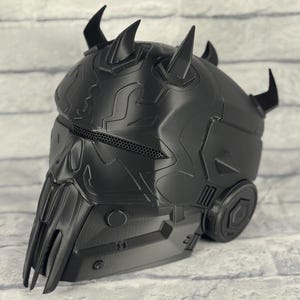 Op de afbeelding: Een zwarte helm met een gedetailleerd, getextureerd oppervlak. De helm heeft meerdere gebogen hoorns en een gezichtsbescherming met een gaasgedeelte. Het ontwerp bevat geometrische vormen en mechanische elementen, wat wijst op een futuristisch of sciencefiction thema.