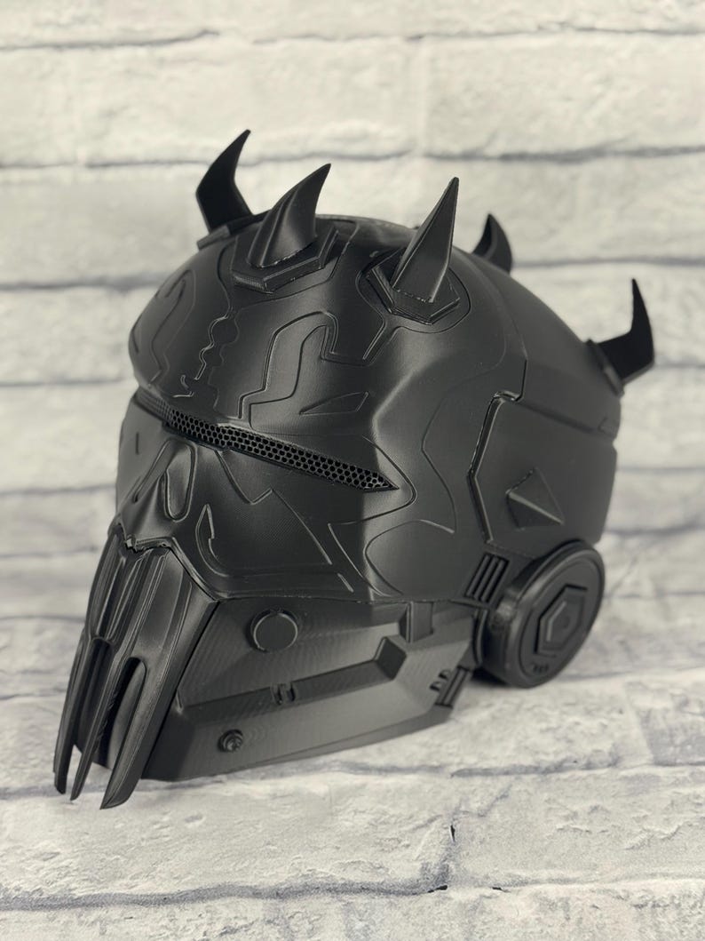 Op de afbeelding: Een zwarte helm met een gedetailleerd, getextureerd oppervlak. De helm heeft meerdere gebogen hoorns en een gezichtsbescherming met een gaasgedeelte. Het ontwerp bevat geometrische vormen en mechanische elementen, wat wijst op een futuristisch of sciencefiction thema.