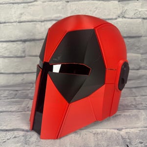 以下が含まれることがあります： 幾何学的なデザインの赤と黒のヘルメット。ヘルメットは滑らかで光沢のある仕上がりで、黒いバイザーエリアが特徴です。ヘルメットの側面には、小さな黒い円形のディテールがあります。ヘルメットはおそらくコスチュームまたはコスプレアイテムです。