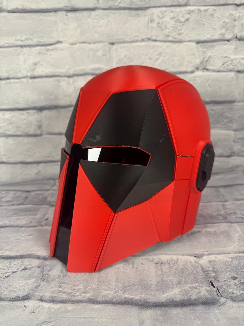 以下が含まれることがあります： 幾何学的なデザインの赤と黒のヘルメット。ヘルメットは滑らかで光沢のある仕上がりで、黒いバイザーエリアが特徴です。ヘルメットの側面には、小さな黒い円形のディテールがあります。ヘルメットはおそらくコスチュームまたはコスプレアイテムです。