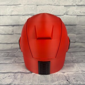 以下が含まれることがあります： 滑らかな仕上がりの鮮やかな赤色のヘルメット。ヘルメットの背面には長方形の黒いディテールがあり、両側には2つの小さな黒いディテールがあります。グレーと白のレンガの壁を背景にしています。