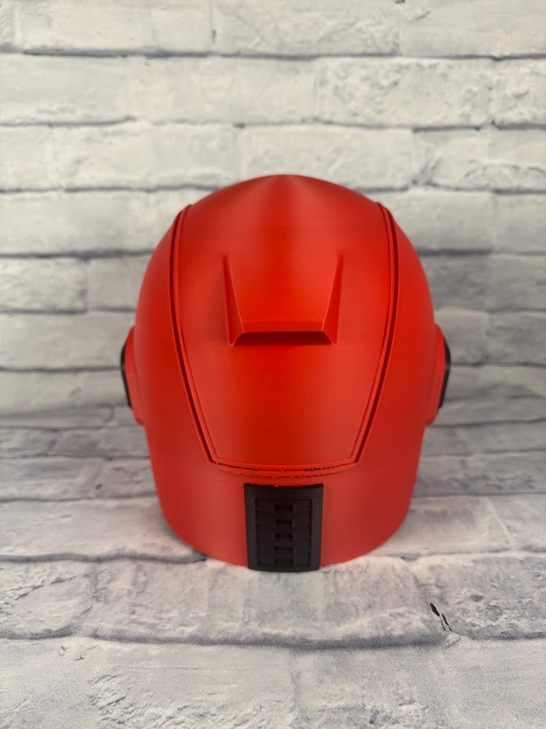 以下が含まれることがあります： 滑らかな仕上がりの鮮やかな赤色のヘルメット。ヘルメットの背面には長方形の黒いディテールがあり、両側には2つの小さな黒いディテールがあります。グレーと白のレンガの壁を背景にしています。