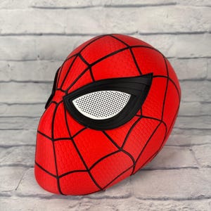 May include: A red full-head mask featuring a black web pattern across the surface. The mask includes large, angular eye openings with black frames and white mesh lenses, designed to cover the entire head with a textured, honeycomb-like finish.