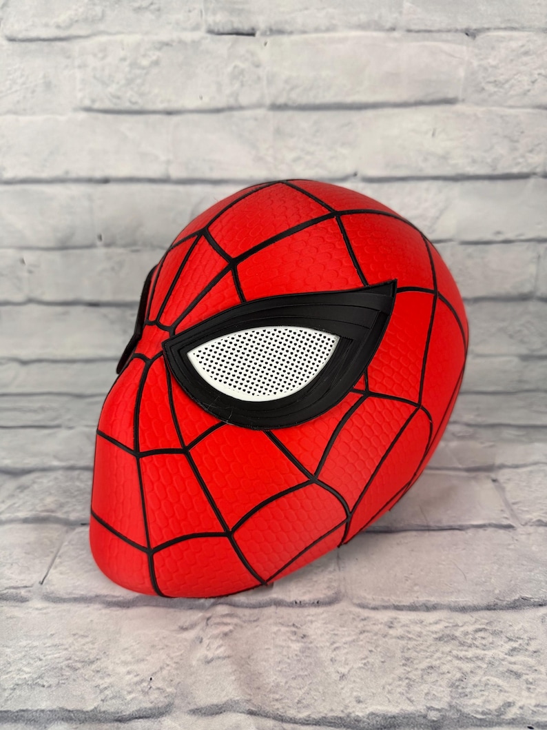 May include: A red full-head mask featuring a black web pattern across the surface. The mask includes large, angular eye openings with black frames and white mesh lenses, designed to cover the entire head with a textured, honeycomb-like finish.