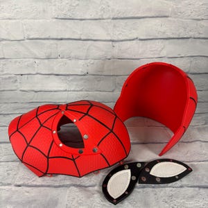 May include: A red and black spider-themed mask kit consisting of a front faceplate with a circular opening, a separate back head shell, and two detachable eye lenses with white mesh inserts. The components feature a textured web pattern.
