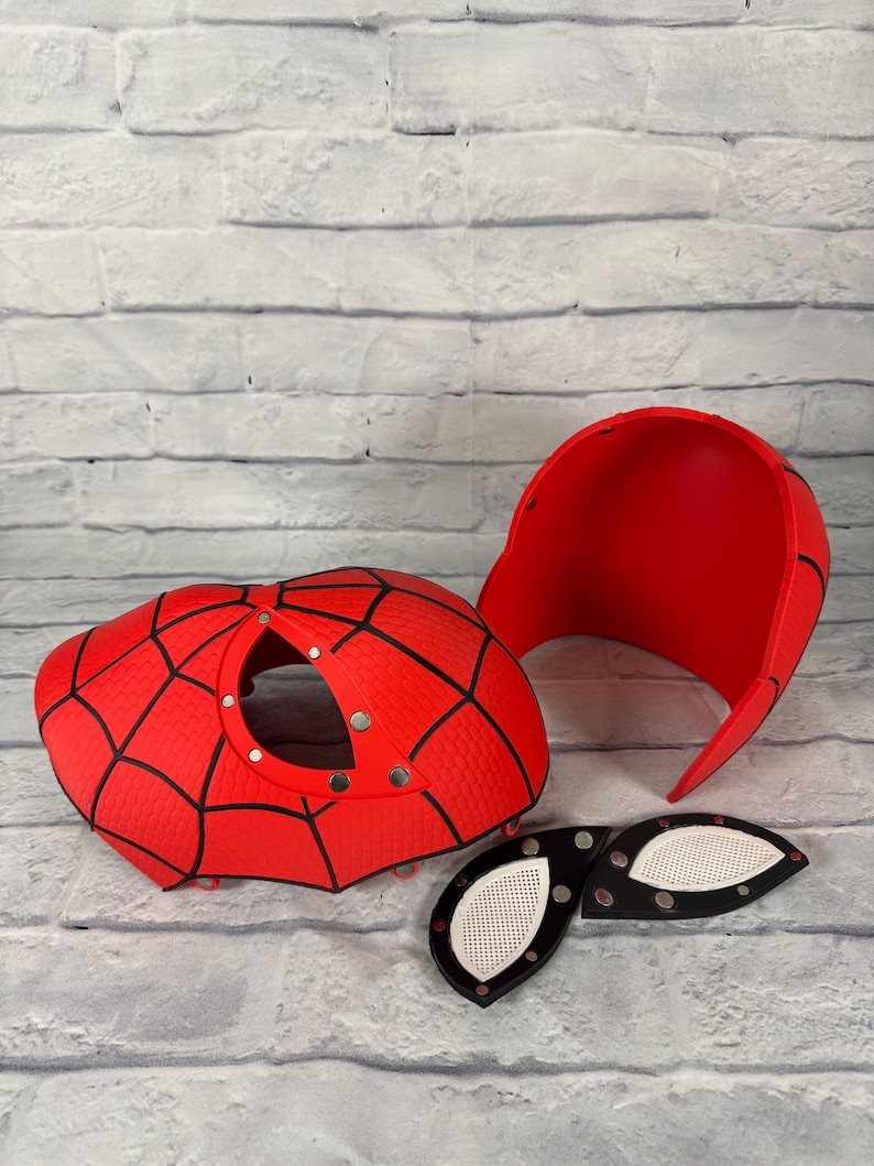 May include: A red and black spider-themed mask kit consisting of a front faceplate with a circular opening, a separate back head shell, and two detachable eye lenses with white mesh inserts. The components feature a textured web pattern.