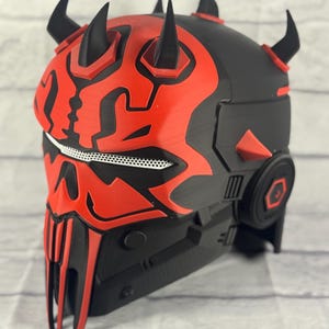 Op de afbeelding: Een zwart en rode helm met een gedetailleerd ontwerp, met prominente hoorns en een kenmerkend gezichtsmasker. De helm heeft een complexe, gelaagde structuur met geometrische vormen en een gaasgedeelte. Het kleurenschema is voornamelijk zwart en rood.