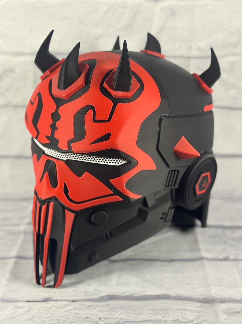 Op de afbeelding: Een zwart en rode helm met een gedetailleerd ontwerp, met prominente hoorns en een kenmerkend gezichtsmasker. De helm heeft een complexe, gelaagde structuur met geometrische vormen en een gaasgedeelte. Het kleurenschema is voornamelijk zwart en rood.
