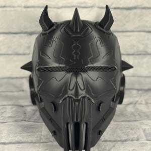Op de afbeelding: Een zwart, ingewikkeld ontworpen helm met een dreigende uitstraling. De helm heeft meerdere spikes en hoorns, een mesh oogbedekking en een gedetailleerd frontpaneel. Het ontwerp suggereert een futuristisch of fantasiethema.