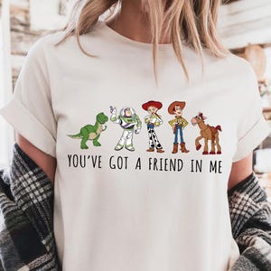 Puede incluir: Camiseta de color marfil con un gr&aacute;fico de personajes de una pel&iacute;cula de animaci&oacute;n popular. El dise&ntilde;o incluye un dinosaurio verde, un guardabosques espacial, dos vaqueros y un caballo, con el texto "YOU'VE GOT A FRIEND IN ME".