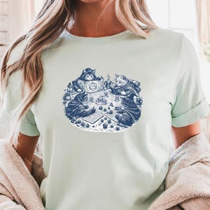 May include: A light green t-shirt featuring a detailed blue illustration of cats playing a tabletop game. The design includes cats wearing cloaks and hats, dice, and game boards. The shirt has short sleeves and a crew neck.