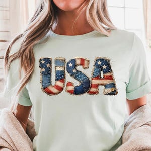 Pode incluir: Uma camiseta verde claro com a palavra "USA" em um design patriótico. As letras são feitas de ganga e apresentam as cores vermelha, branca e azul da bandeira americana. As mangas estão enroladas.