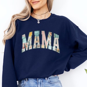 May include: Navy blue sweatshirt with the word "MAMA" in a colorful, patchwork-style design across the chest. The sweatshirt has long sleeves and a crew neck. The word "Navy" is in a circle in the upper right corner.