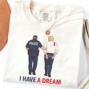 Può includere: T-shirt Comfort Colors color avorio con un'illustrazione a cartoni animati di un poliziotto che scorta un uomo ammanettato. Il testo "I HAVE A DREAM" è stampato in rosso e nero in basso.