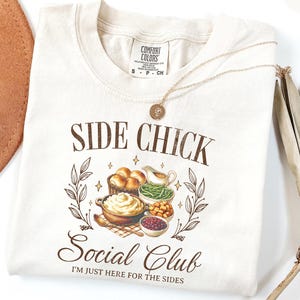 May include: Ivory-colored t-shirt with the text "SIDE CHICK Social Club" and the phrase "I'M JUST HERE FOR THE SIDES". The graphic features illustrations of Thanksgiving side dishes. A gold necklace is draped on the shirt.