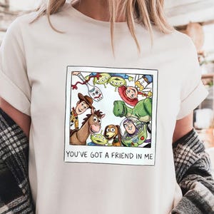 Op de afbeelding: Ivoorkleurig T-shirt met een ingelijste cartoonafbeelding van Toy Story-personages, waaronder Woody en Buzz Lightyear. De tekst "YOU'VE GOT A FRIEND IN ME" staat onder de afbeelding. Het T-shirt is licht cr&egrave;mekleurig.