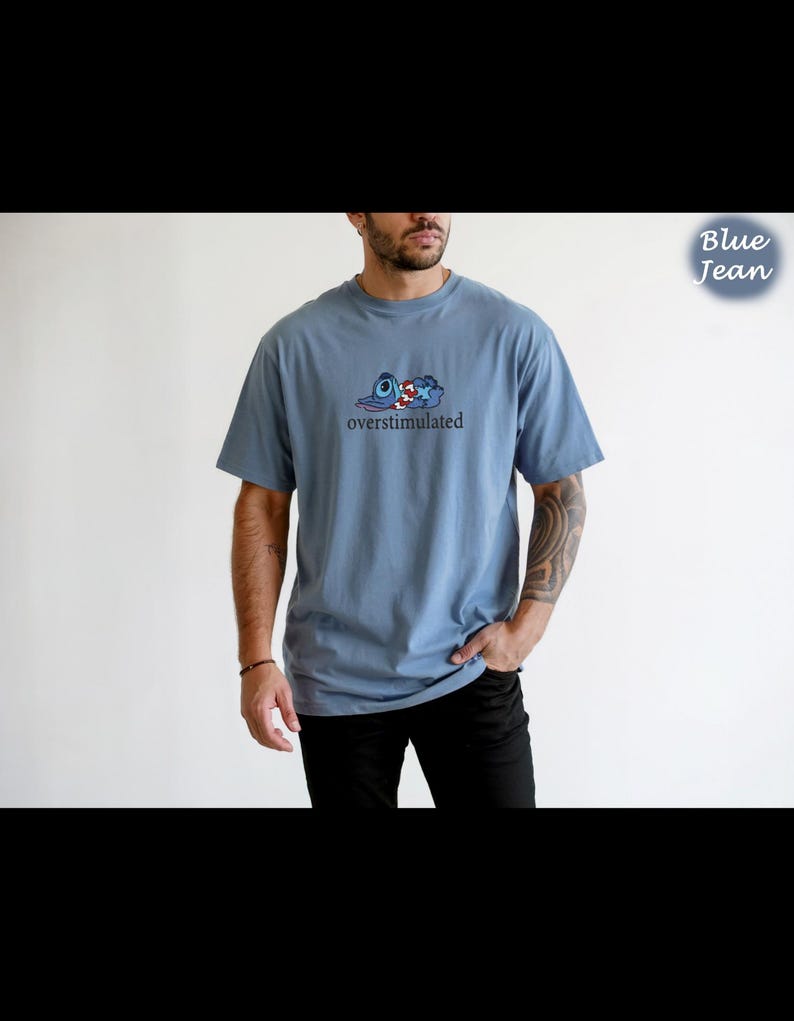 以下が含まれることがあります： 「overstimulated」の黒い文字が入った、青色の半袖Tシャツ。青、赤、白の漫画のグラフィックが特徴です。「Blue Jean」の文字が右上隅に青色で表示されています。