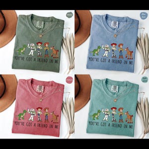 Puede incluir: Cuatro camisetas en verde musgo, azul vaquero, carmes&iacute; y verde claro, cada una con un gr&aacute;fico de personajes de dibujos animados y el texto "YOU'VE GOT A FRIEND IN ME." Cada camiseta tiene un collar.
