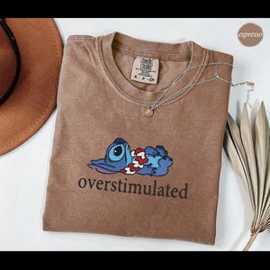 以下が含まれることがあります： エスプレッソ色のComfort Colors Tシャツ。漫画のキャラクターと「overstimulated」の文字が黒でプリントされています。シルバーのペンダントネックレスがシャツの上にあります。クルーネック、半袖です。