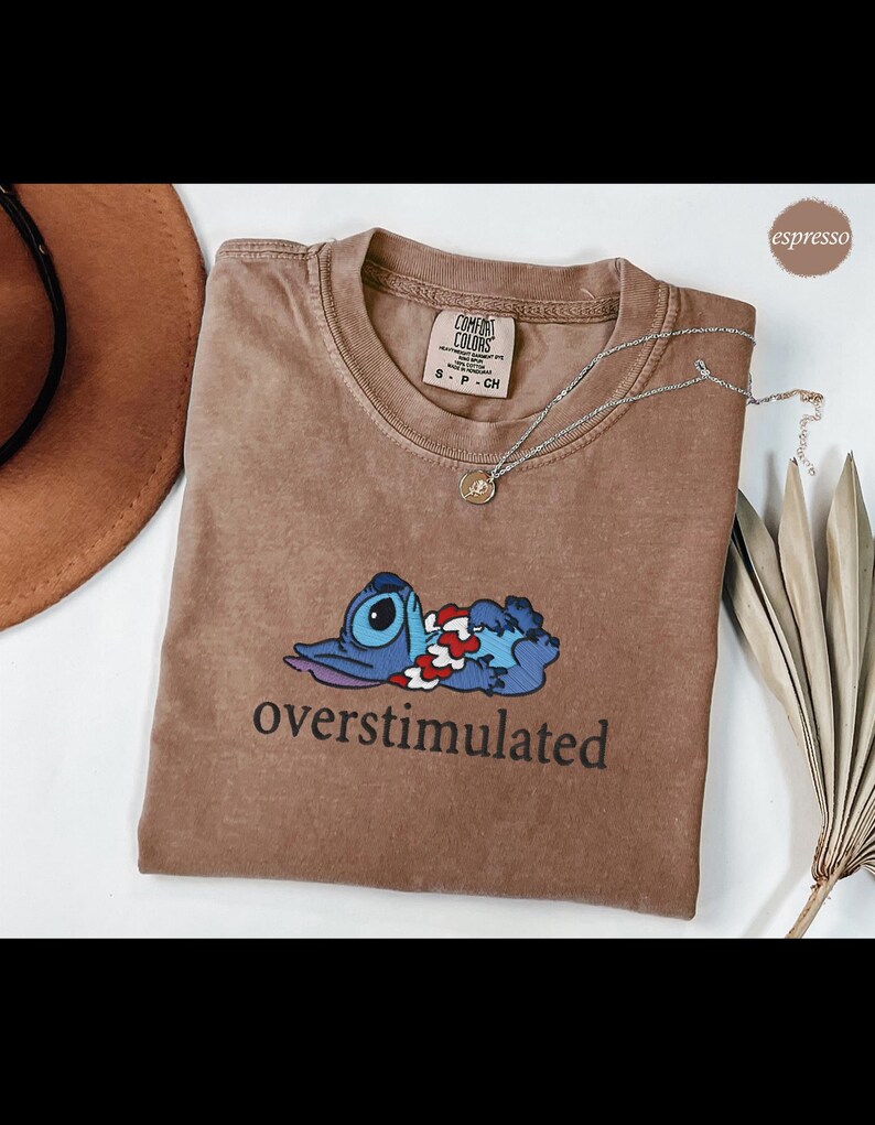 以下が含まれることがあります： エスプレッソ色のComfort Colors Tシャツ。漫画のキャラクターと「overstimulated」の文字が黒でプリントされています。シルバーのペンダントネックレスがシャツの上にあります。クルーネック、半袖です。
