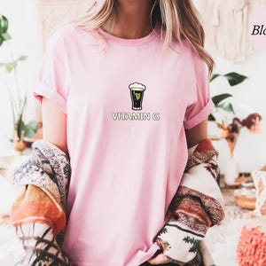 May include: A light pink t-shirt with rolled-up sleeves featuring a graphic of a pint of stout with the text "VITAMIN G" below. The shirt is part of a clothing collection.