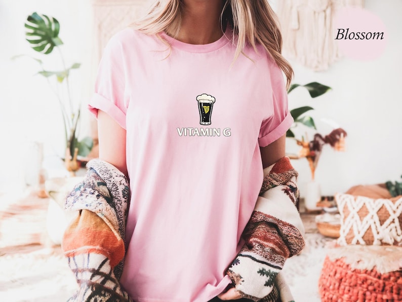May include: A light pink t-shirt with rolled-up sleeves featuring a graphic of a pint of stout with the text "VITAMIN G" below. The shirt is part of a clothing collection.