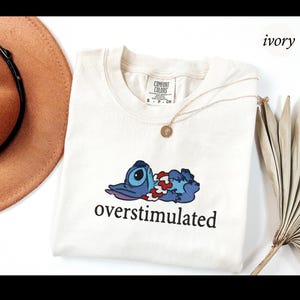 以下が含まれることがあります： アイボリー色のTシャツ。青い漫画のキャラクターと「overstimulated」の文字がプリントされています。Tシャツはクルーネックで半袖です。ペンダント付きのゴールドネックレスも確認できます。