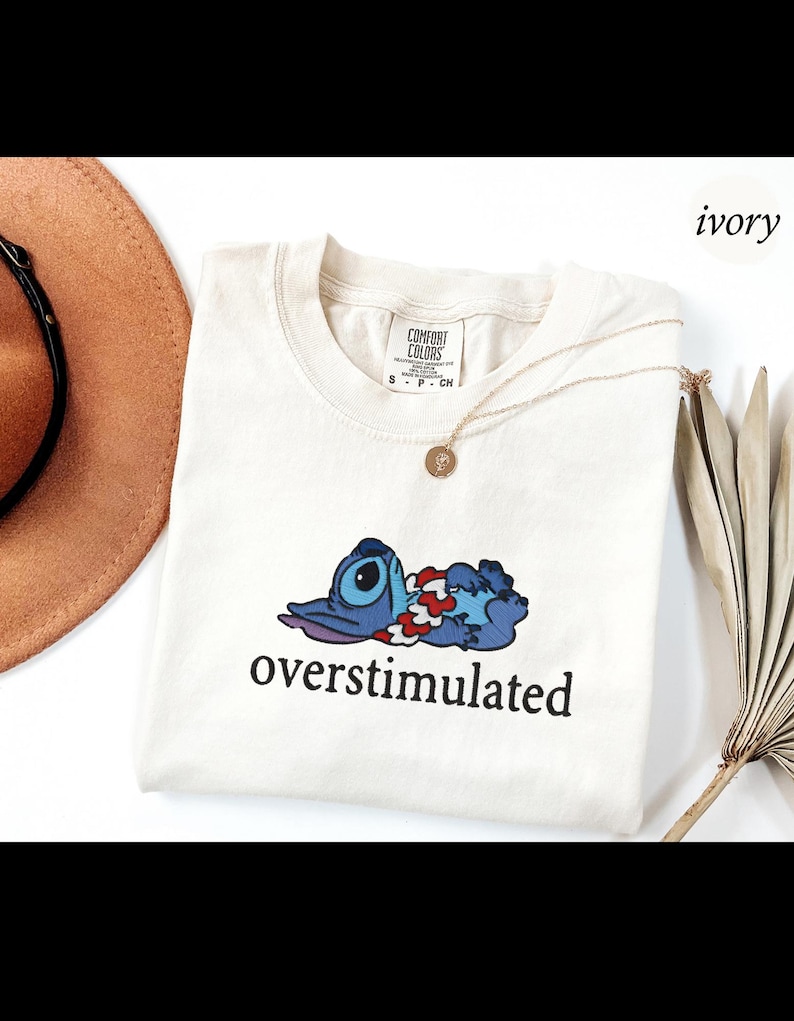 以下が含まれることがあります： アイボリー色のTシャツ。青い漫画のキャラクターと「overstimulated」の文字がプリントされています。Tシャツはクルーネックで半袖です。ペンダント付きのゴールドネックレスも確認できます。